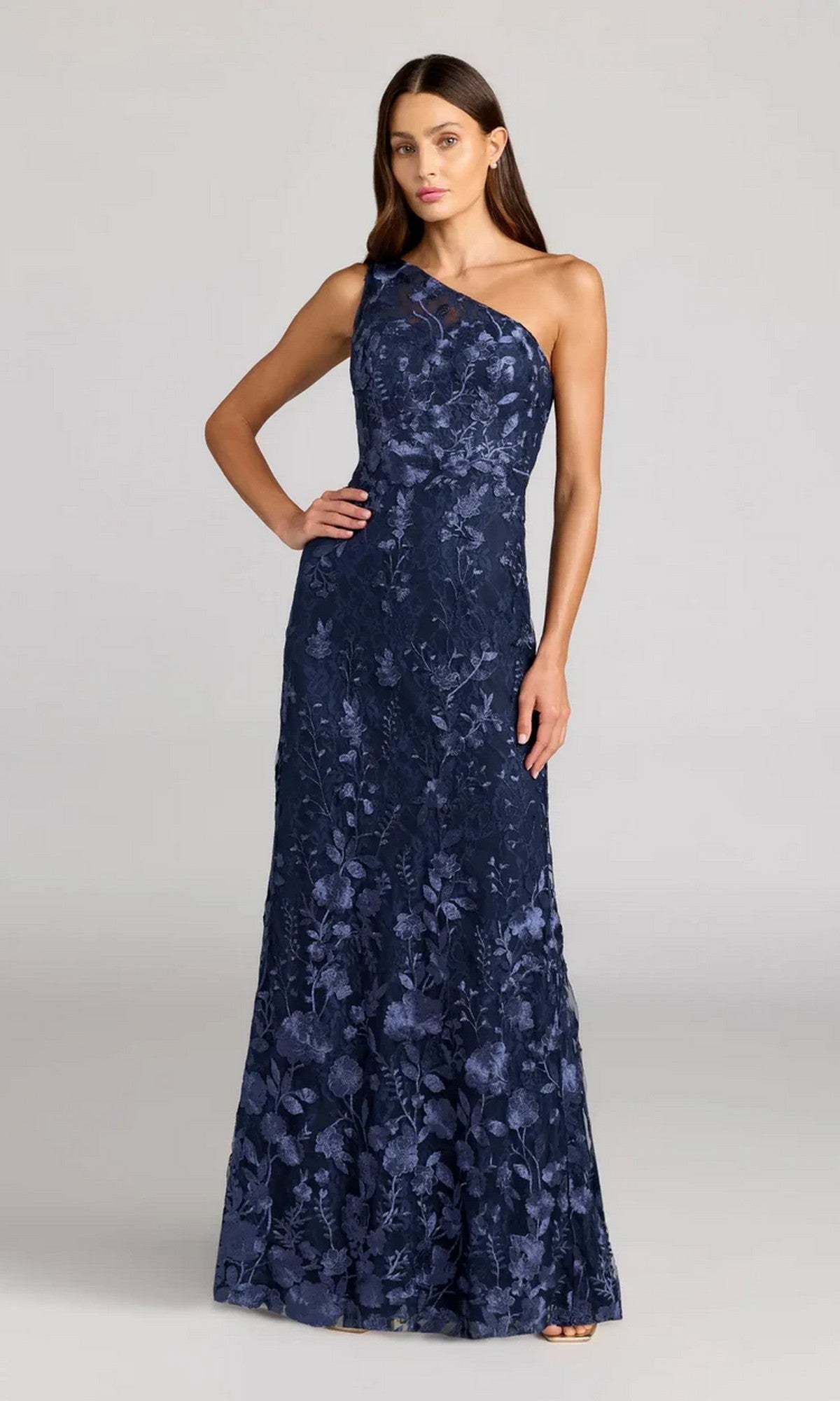 A woman stands against a light background wearing the Mac Duggal 68606 Long Floral-Print Formal Dress in Midnight Multi—a floor-length, one-shoulder gown with an elegant floral pattern.